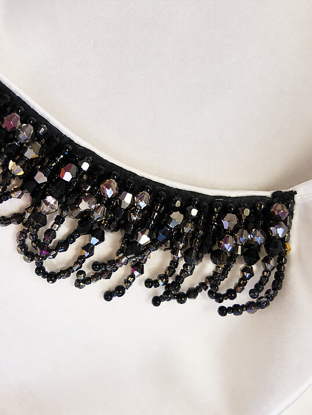 A close-up of a black beaded tiara or crown with faceted black and iridescent beads, resting on a white fabric background.