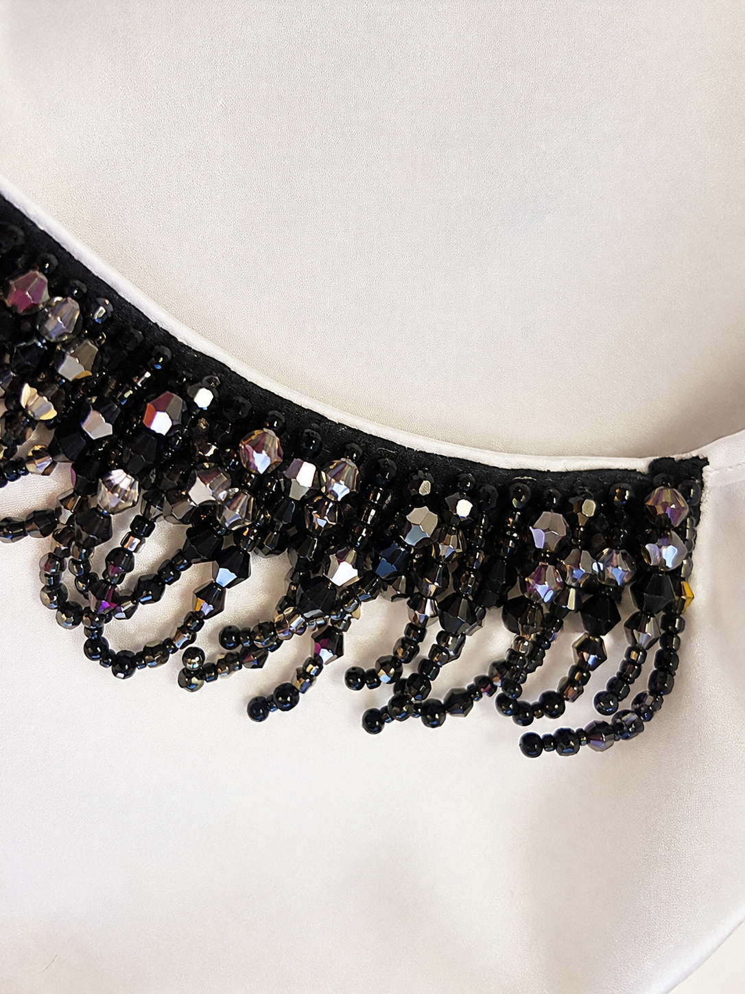 A close-up of a black beaded tiara or crown with faceted black and iridescent beads, resting on a white fabric background.